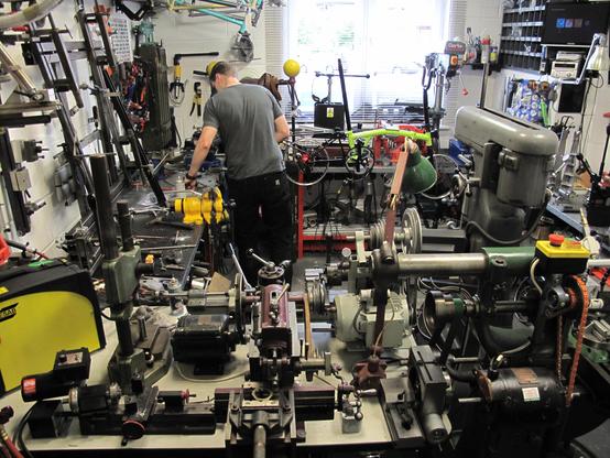 Eine sehr enge Werkstatt voller alter Metallbearbeitungsmaschinen, Rohrbieg-Gerätschaften und Schablonen zum Löten. Zum Fenster hin ist ein Brompton Faltrad zu sehen, das sich noch im Aufbau befindet. Daneben arbeitet ein Mann im grauen T-Shirt mit dem Rücken zum Fotografen.