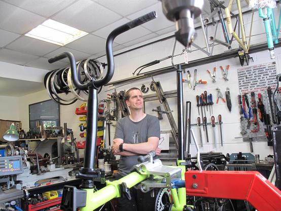 Ein Mann im grauen T-Shirt steht mit verschränkten Armen in seiner Fahrradwerkstatt und unterhält sich mit dem Fotografen. Dabei schaut er am Fotografen vorbei zum Fenster hinaus. Im Vordergrund ein halb aufgebautes Brompton Faltrad in einem Montageständer. An der Wand im Hintergrund zahlreiche Werkzeuge. An der Decke hängen Fahrradrahmen.