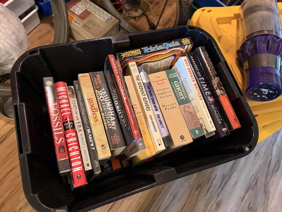 A black plastic storage bin is filled upright with paperback and hardcover books, showing their spines with various titles and colors, sitting on a wooden floor in a cluttered room; surrounding the bin are other household items including stacked boxes in the background and a yellow-lidded container with a purple cordless vacuum canister resting on top at the right edge of the image.