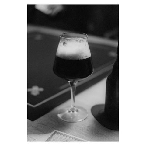 Ein schwarz-weiß Foto. Mittig ein Bierglas mit dunklem Bier. Rechts davon eine Bügelbierflasche. Im Vordergrund liegen Spiekarten auf einem Tisch. 