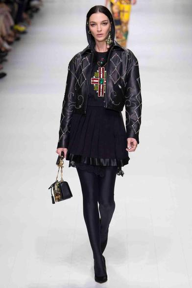 A runway model in a black leather jacket embellished with crystals, a black t-shirt featuring a jewel-encrusted cross design, a pleated black skirt, black opaque tights, and black pointed-toe heels, holding a small black jeweled handbag. 