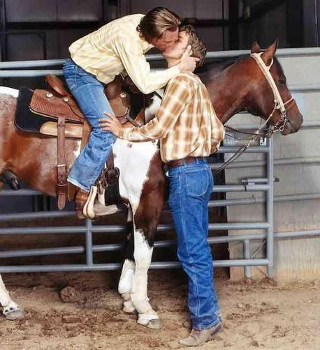 Two men kissing, one seated on a saddled horse, the other standing beside the horse, both in western attire.