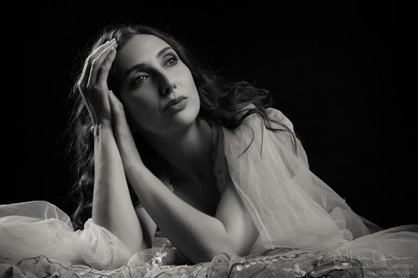 Black and white photo of model Anoush Anou, head and shoulders. She is leaning on a table top, surrounded by sheer material. Her gaze is looking up and off to camera right. The entire scene is low key with heavy shadows, giving a film noir look.