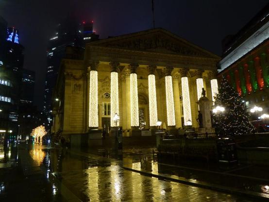 Classical building with eight pillars, illuminated with many golden lights, reflected in a wet London street at night.