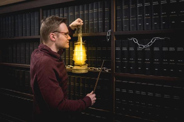 A young wizard wearing glasses stands in front of a bookshelf filled entirely with black books and adorned with chains. In his left hand, he holds an old oil lamp that truly illuminates the gloomy scene, and in his right hand, he holds a magic wand.