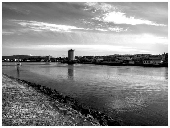 A high contrast, black and white photograph of the Mersey River in Devonport, Tasmania. In the foreground, a grassy bank slopes down to a rocky seawall at the edge of the dark, reflective river.

Dominating the middle ground, a tall, cylindrical building, possibly a silo or water tower, stands near the river's edge. Buildings line the distant bank, backed by low hills under a dramatically clouded sky.