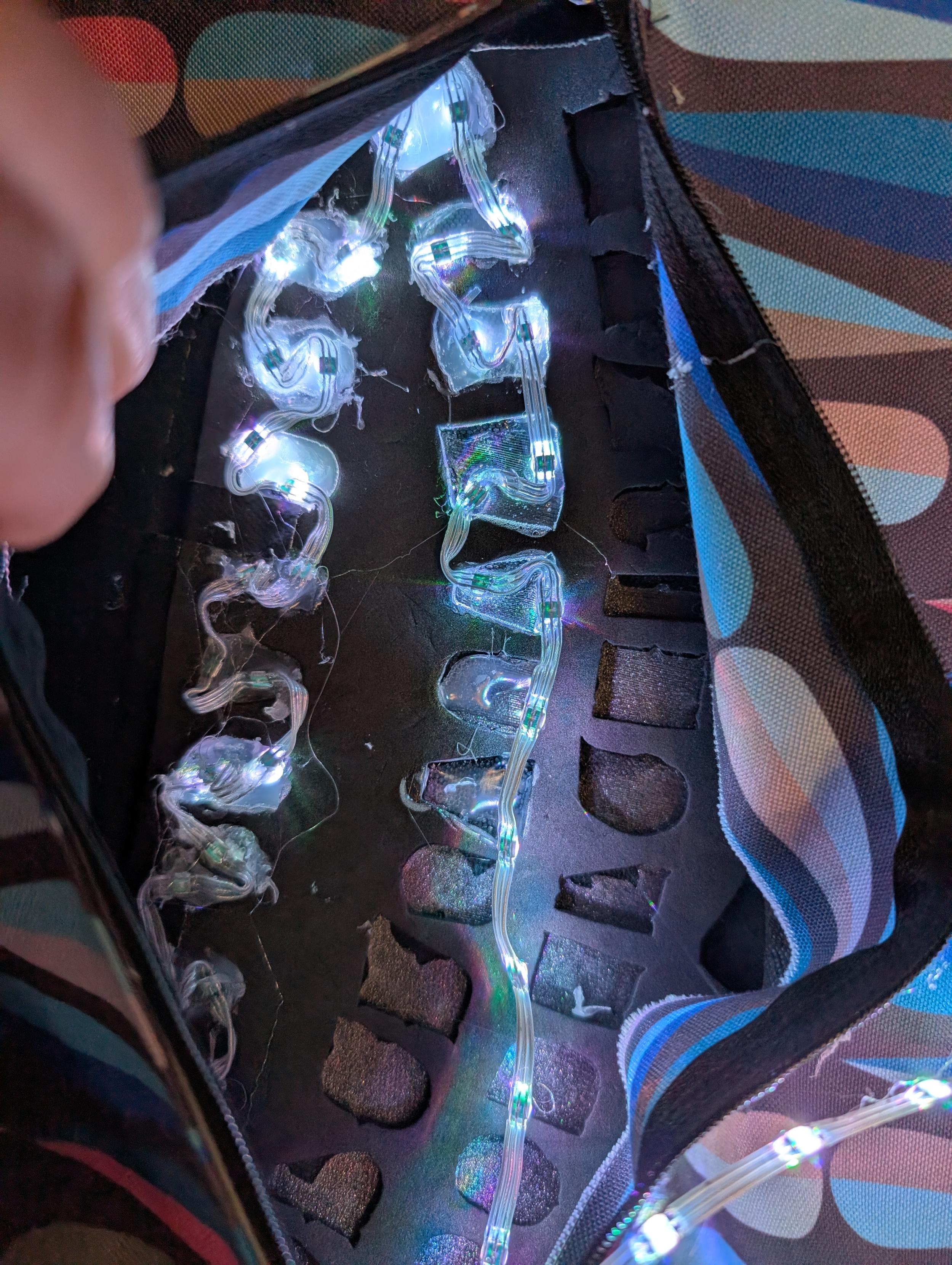 The view inside the front flap of the messenger bag with a panel zipped open to reveal the back side of the illuminated letters which are surrounded by EVA foam with cutouts at the letter positions. Each letter cutout is filled with hot glues and has two LEDs from an LED string attached to eliminate the letter from behind.