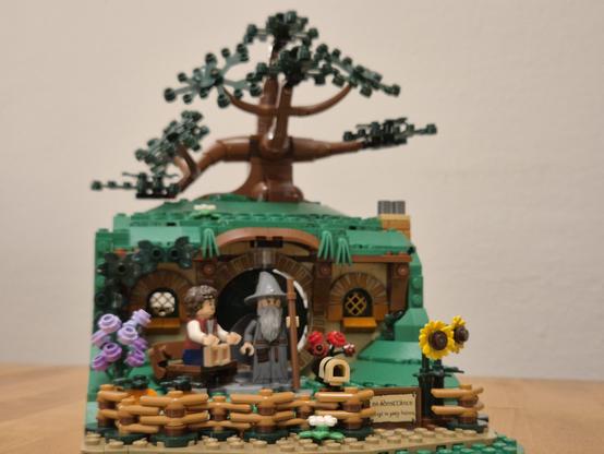 This image showcases a beautifully crafted LEGO diorama inspired by a fantasy setting, reminiscent of a hobbit hole from a beloved series. The scene features a cozy hobbit house built into a hill, complete with a large tree growing above it.

Key elements of the diorama include:

- **Hobbit Hole:** The round green door is a signature feature of the hobbit hole, surrounded by a detailed facade with arched windows and earthy tones. The door is framed by lush, green LEGO bricks, giving it a natural, built-into-the-hill look.

- **Tree:** A large, detailed tree with sprawling branches grows above the hobbit hole, adding a sense of depth and realism to the scene.

- **Minifigures:** Two LEGO minifigures are positioned in front of the hobbit hole. One minifigure, dressed in a vest with curly brown hair, is holding a box. The other minifigure, with a long beard and a wizard-like appearance, is holding a staff, adding a magical touch to the scene.

- **Garden and Decorations:** The area around the hobbit hole is adorned with various elements:
  - A wooden fence with a small gate.
  - Colorful flowers, including purple and red blooms, and sunflowers, adding vibrancy to the scene.
  - A small sign that reads "No Admittance Except on Party Business," a fun nod to the fantasy theme.
  - A small wheelbarrow and a birdhouse, enhancing the cozy and lived-in feel of the setting.