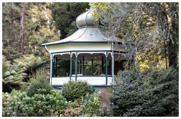 A charming white and green rotunda structure, featuring ornate pillars and a curved, dome like roof, nestled deeply within a lush, temperate garden or park setting.

The rotunda is partially obscured by dense green foliage and trees with dappled sunlight filtering through the canopy.