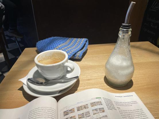 A coffee cup on a saucer with a spoon rests on a wooden table, accompanied by a glass bottle with a straw and a partially open book displaying text and images. A blue and tan knitted item is positioned next to the coffee.