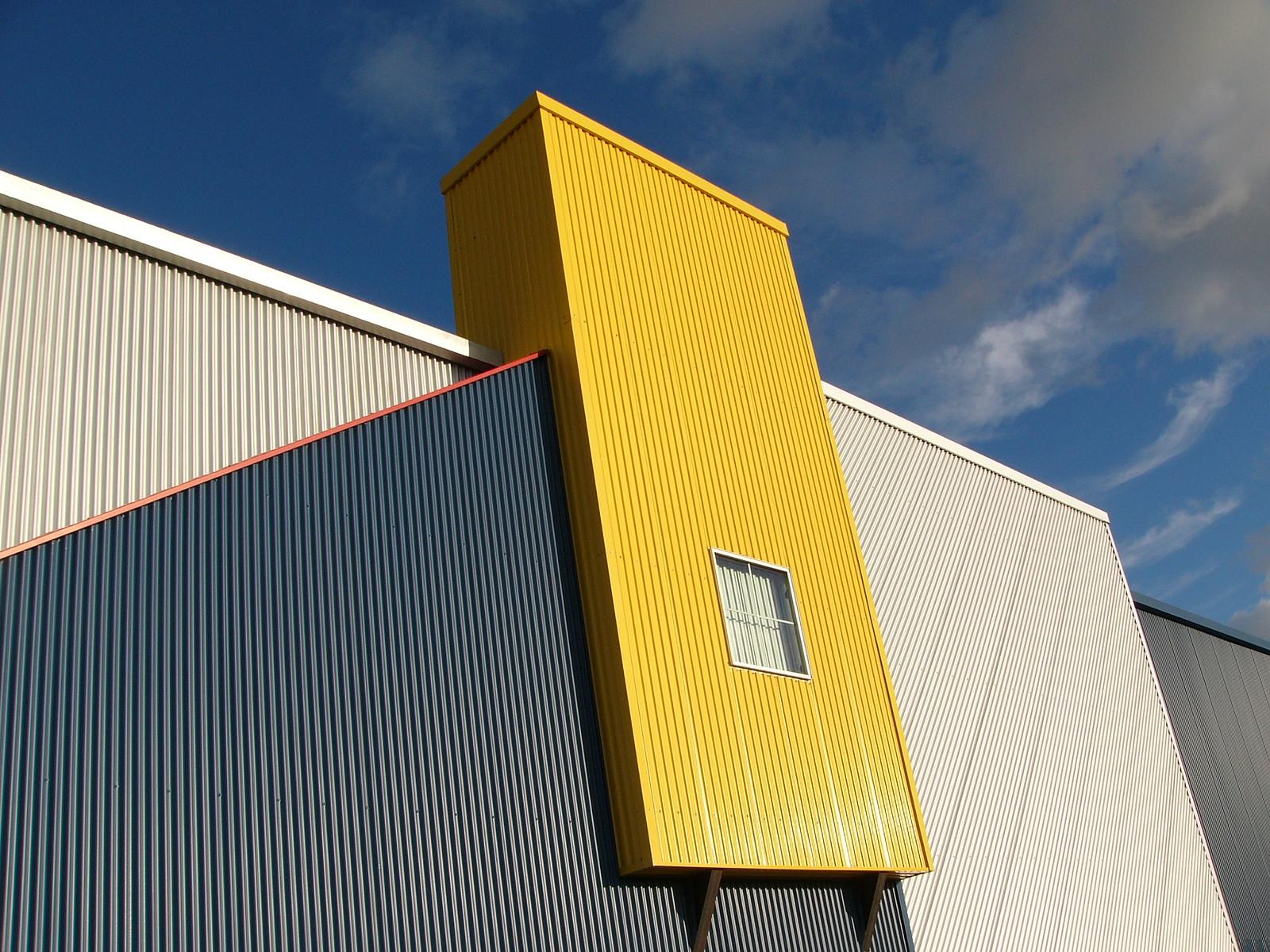 a single window on a weirdly shaped building with corrugated sides painted white, gray, and yellow, under a blue sky