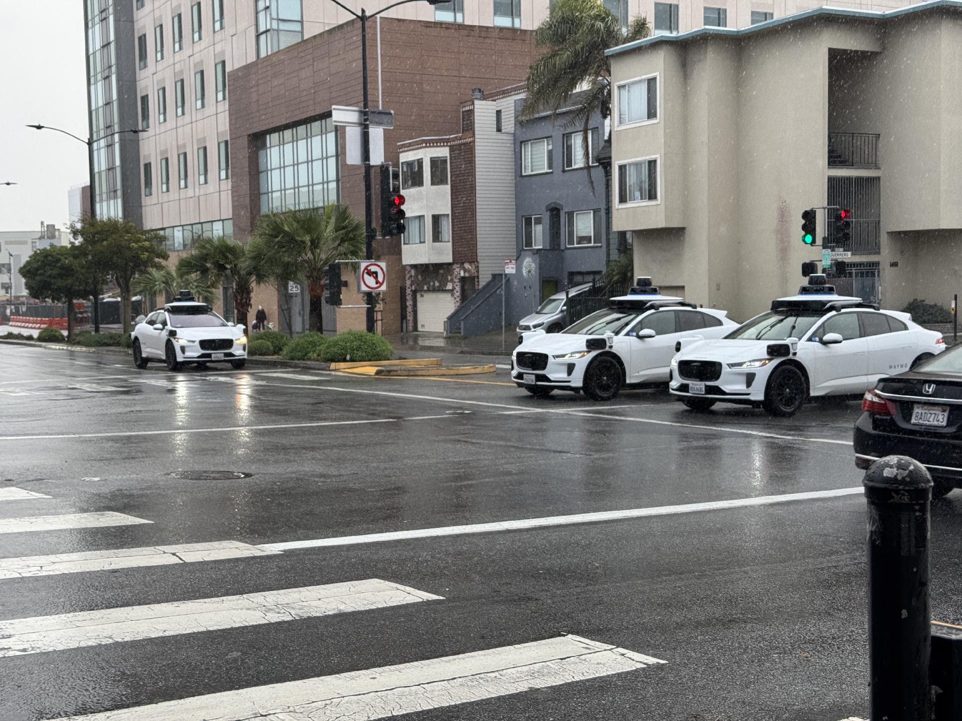 Three white Waymos (autonomous vehicles) at an intersection of Cesar Chavez and Guerrero. It’s a rainy day. No passengers are visible, but the windows are tinted.