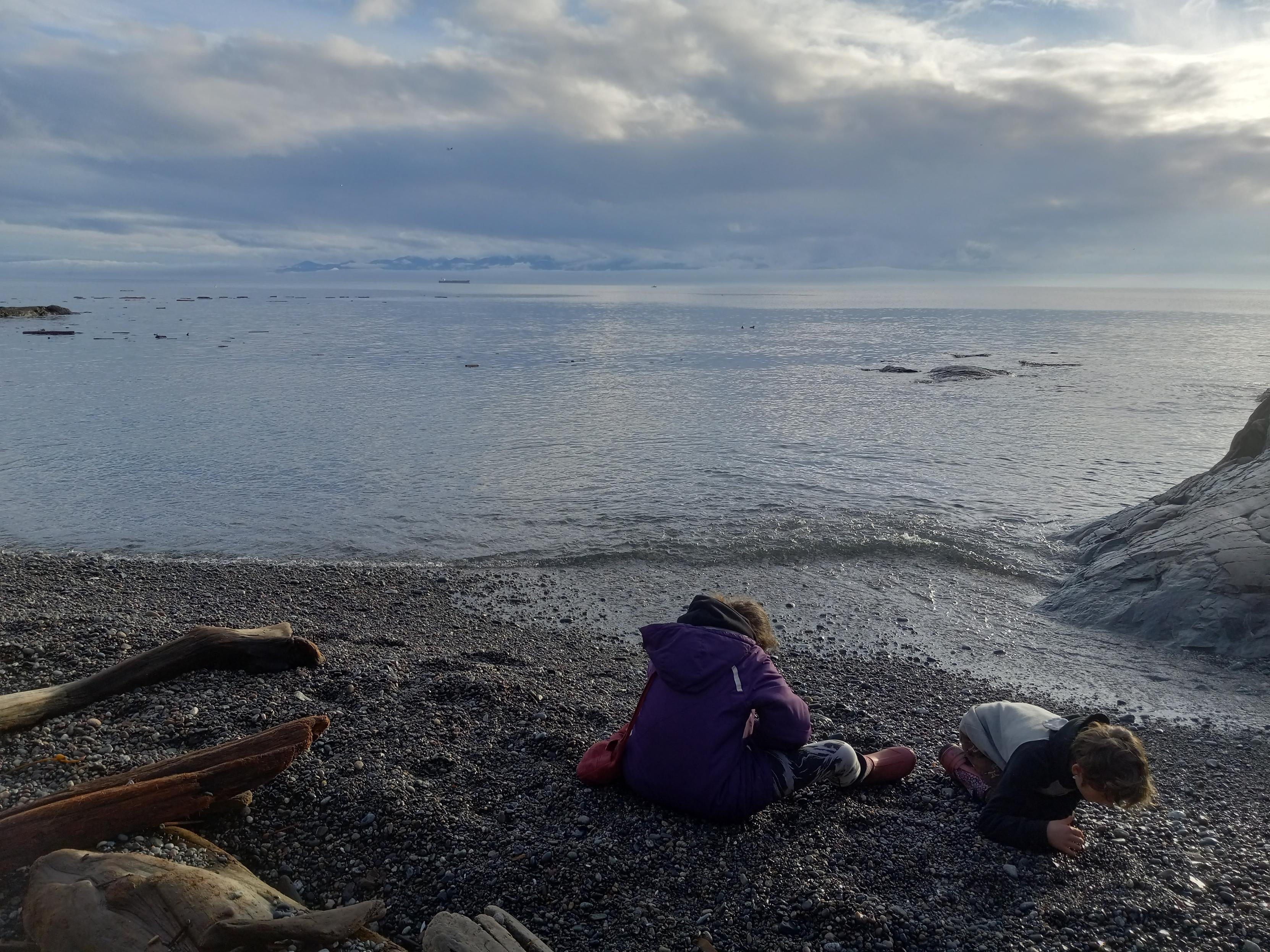 2 kids sitting on a beach made of small rocks, intensely playing with the rocks around them.  There's some driftwood and seaweed laying on the beach, and small waves hitting the rocky beach.  In the calm ocean behind the kids, there's some floating driftwood, and far in the distance, a large ship.  Beyond that are some really beautiful flowing clouds, with the snow-covered Olympic mountains peeking through.