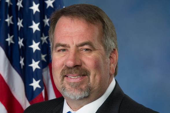 Man with short brown hair and a trimmed goatee poses for a formal portrait in a suit and tie, smiling slightly, with a U.S. flag and blue backdrop behind him.