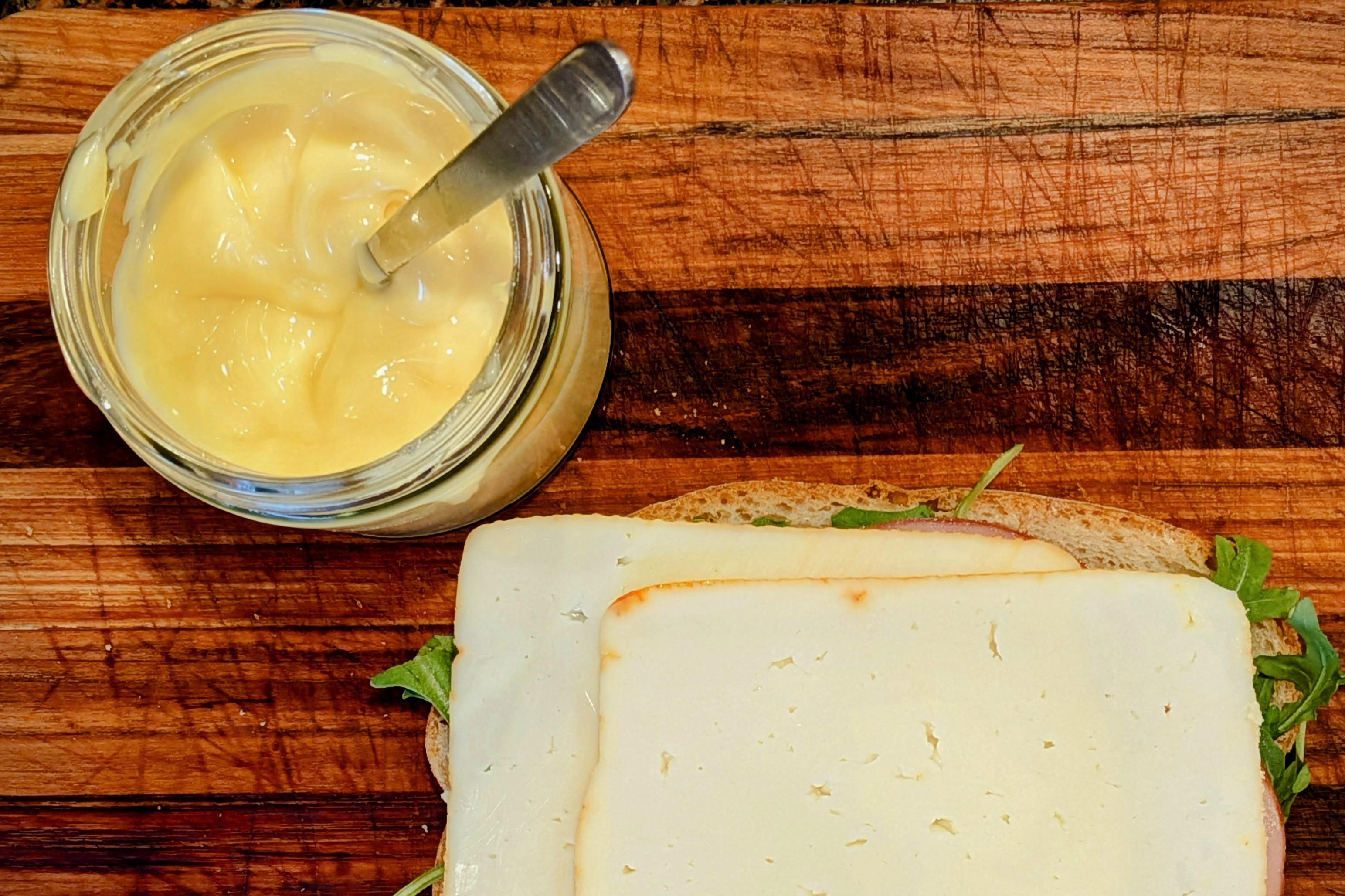 A vanity shot from above of a teak cutting board with the bottom half of a sandwich covered with cheese slices and a small mason jar of a yellow glistening mayonnaise standing next to it.