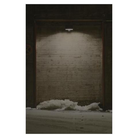 Frontaler Blick auf eine Betonwand. Mit Stahlträgern eingerahmt ist ein rechteckiger Teil der Wand um ein paar centimeter zurück gesetzt. Eine mittig auf der Wand montierte Leuchte strahlt die Wand von oben nach unten aus, wodurch diese wie eingerahmt wirkt. Auf dem Boden vor der Wand liegt ein kleiner Schneehaufen. 