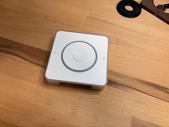A white, square-shaped device with a circular button in the center placed on a wooden surface.