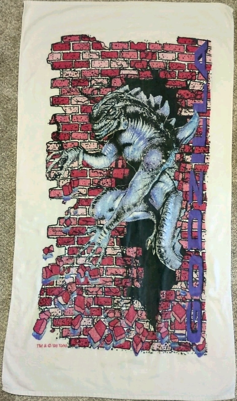A photo of a towel with a weird, thin, humanoid lizard-monster bursting through a brick walk. Text says "Godzilla" but this weird monster doesn't look like Godzilla.