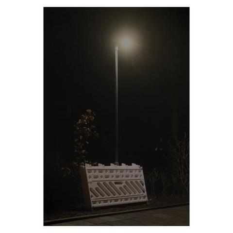Foto einer nächtlichen Straßenszene. Eine Straßenlaterne steht mittig im Bild an einem Bürgersteig. Weiße Absperrgitter einer Baustelle lehnen hintereinander gestapelt an der Laterne. Der Hintergrund ist dunkel. 