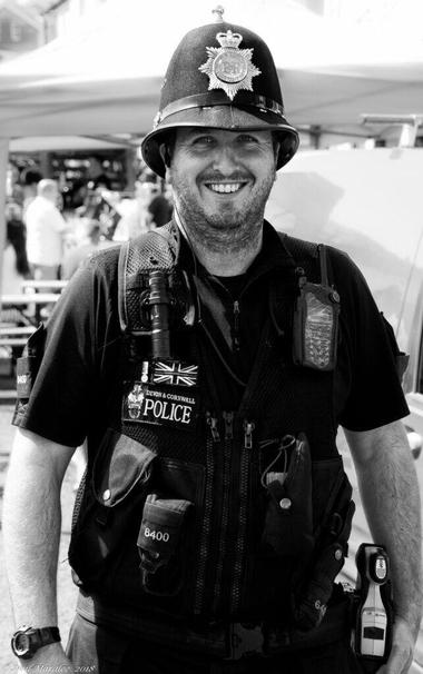 A black-and-white photograph of a smiling British police officer wearing a helmet and multiple items of equipment around his body.