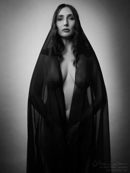 Model Anoush Anou poses standing nude, draped with a sheer black fabric that she holds closed like a robe. She engages the viewer.