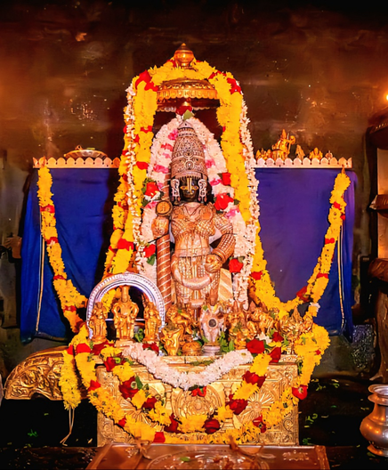 Idol of Hindu deity Krishna standing on a gold-covered pedestal, clad in diamond-studded armor, holding a similarly-clad staff in right hand, left hold on front left waist; a small gold umbrella overhead. A string of white flowers (some bands of red flowers) around Krishna. A string of yellow flowers and a thin string of white flowers flow down from the top of the umbrella onto both sides of Krishna, all the way to the pedestal. A string of yellow flowers hangs curved in front of the pedestal. Several other small idols at Krishna's feet. Dark blue cloth background. A long string of yellow flowers hangs from one end of the background to the other.