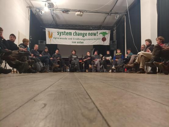 50 Menschen sitzen in einem Saal im Kreis. Im Hintergrund das Banner "System Change now! Ernährungssouveränität und Agrarwende von unten!" 