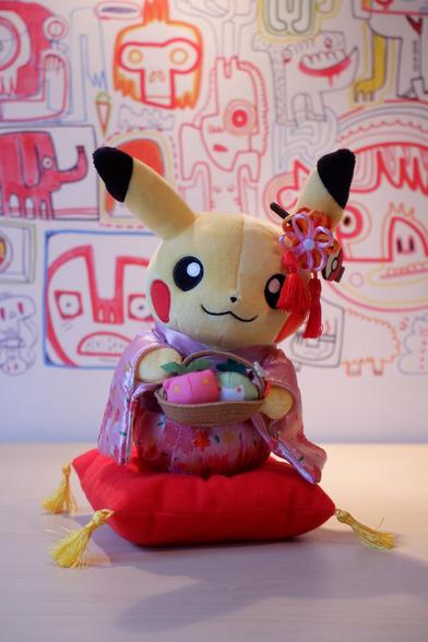 A plush Pikachu toy dressed in traditional Japanese attire, holding a basket with pink and white fabric items, sitting on a red cushion.