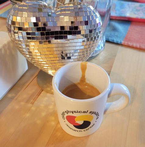 Daytime interior photo of a white, 3 ounce, ceramic espresso (demitasse) cup with a shot of espresso, on a pine-wood surface, in front of a 4-inch decorative heart covered in silver reflective sequins. The blue text on the cup reads "Cyber-Physical Systems Research Center" and wraps in a circle around the logo, which looks like a loosely interlaced, rounded triquetra with thick knots in solid red, yellow, and blue (each its own color).