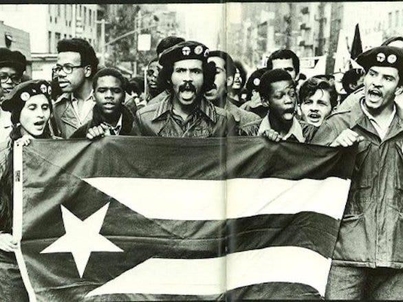 A group of Black Americans and Puerto Ricans marching. They're holding up a big Puerto Rican flag. 