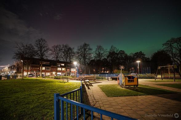 Nachtfotoโs. Zicht op speeltuin en huizen, met groenig en roze lucht, wat wolkjes.