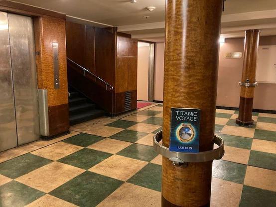 Titanic Voyage sitting on a railing at a wooden column on the queen Mary. An elevator is in view, with original turquoise-and-white checked linoleum flooring.