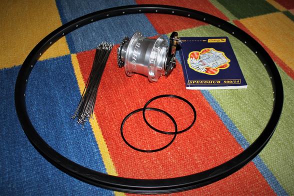 Auf einem bunten Teppich liegen der Felgenring Ryde Andra 30 schwarz, die 38 DT-Swiss Speichen silber, das Rohloff Handbuch, die 2 Flanschringe und die Rohloff Speedhub in silber.