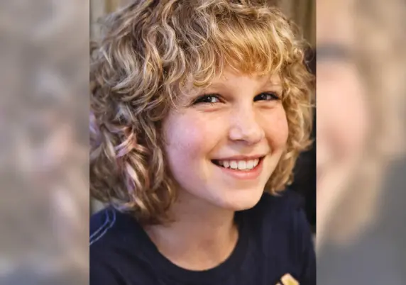 A close-up portrait of a smiling child with curly blond hair and freckles, wearing a dark shirt, looking slightly off-camera with a warm, cheerful expression against a softly blurred background.