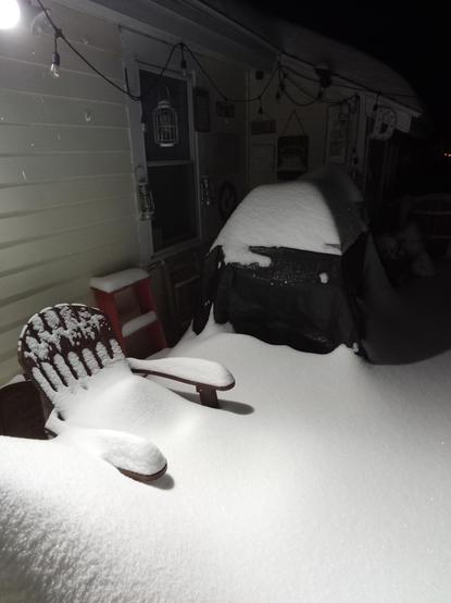 An outdoor deck with Adirondack chair and a small bar covered in a tarp and piled with snow. The seat of the chair can no longer be seen. The snow comes nearly up to the arm rests. And it's still snowing. 