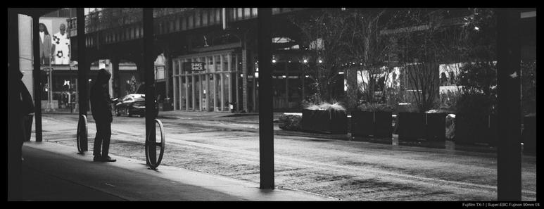 A person stands beneath scaffolding, sandwiched between circular bike racks and structural pillars, as they look at their phone.