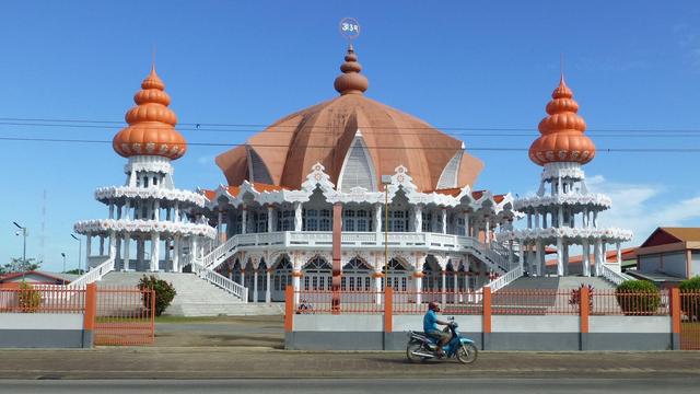 an image of a light orange-red domed building, two spired pavilions on the left and right, the large domes look like large blooming flowers, the walls white , the sky blue behind, on the road in front is a man on a motorcycle parked 