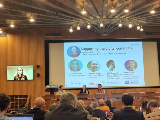 Panel with 3 people onstage and one online, discussing "supporting the digital commons".