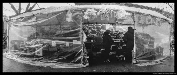 A shopper looks at produce inside of a partially-enclosed street stall, standing alongside the shopkeeper. Both are bundled in heavy coats to stave off cold weather.