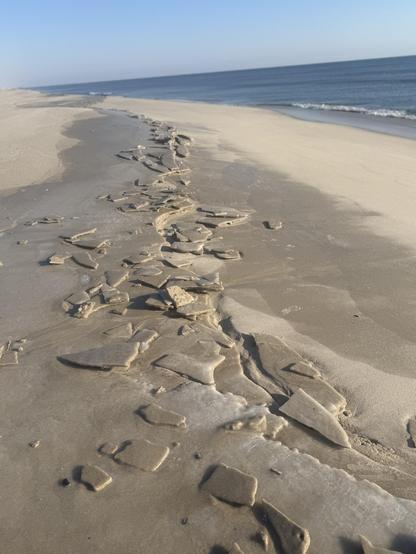 The left of the frame is the beach.  Where the waves come up and retreat, the sand has ben frozen into many odd geometric shapes. The upper right of the frame is an extremely calm Atlantic Ocean and the pale, cold blue sky