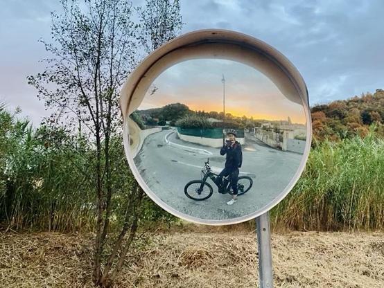 Une cycliste à l’arrêt sur son VTT électrique, reflétée dans un miroir convexe. L'arrière-plan montre une route en épingle à cheveux, de la verdure et le ciel au coucher du soleil. Le centre de la photo est ainsi orangé alors que le reste du ciel est gris de nuages. 