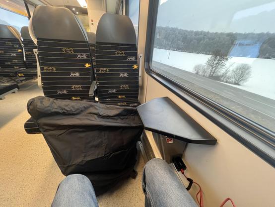 Vierersitzplatz in einem Nahverkehrszug. Vor den zwei Sitzen mit Rücken zur Fahrtrichtung steht ein zusammengefaltetes Faltrad mit einer schwarzen Hülle darüber. 

Am Fenster ein kleines Tischchen, darunter eine Steckdose, in der ein Handyladegerät steckt. Vor dem Fenster schneebedeckte Landschaft bei trübem Wetter. 

Im Vordergrund zwei Knie in Jeans