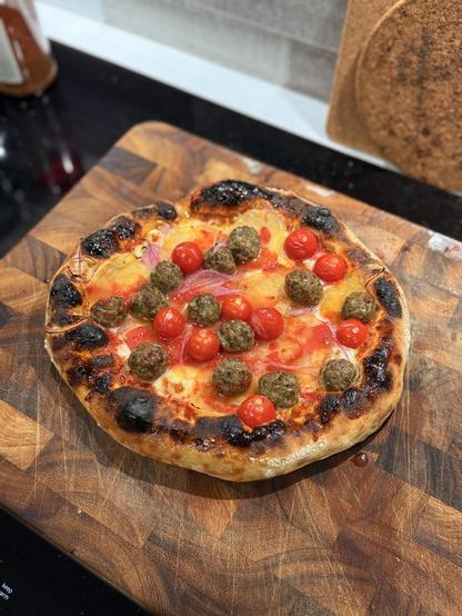 A homemade pizza with a charred crust, topped with cherry tomatoes and kefta meatballs, sits on a wooden cutting board. The pizza has sauce on the bottom, then mozzarella, garlic, sliced red onions, thinly sliced fermented potatoes, and the kefta and tomatoes. 