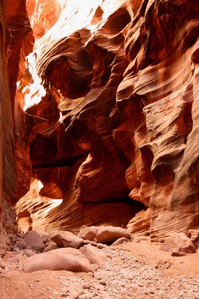 Photo looking up into a corner of a steep-walled desert slot canyon of wavy textured red rock. The bottom of the canyon is red sand and pebbles.