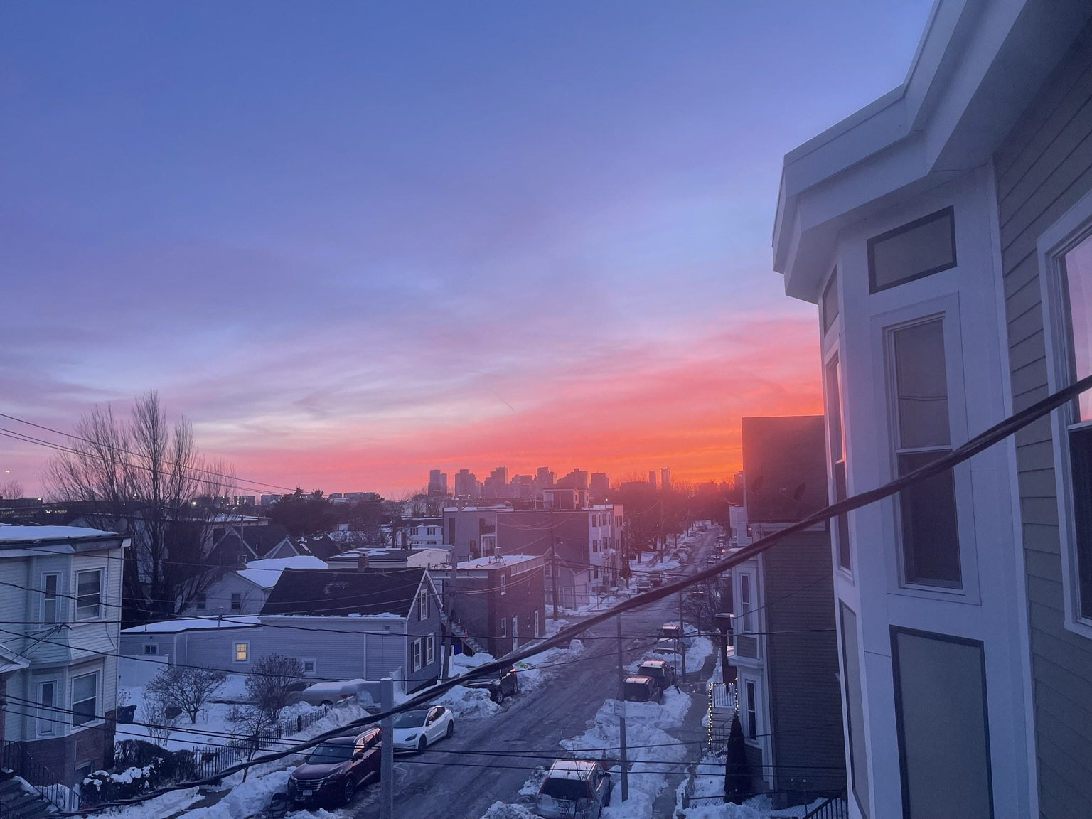 This is an expanded view, looking down my street to the city of Austin about 5 miles away. You can see the sky starting to change colors halfway through the photo. At the top is a grayish blue hit by clouds, followed by pinks reds and oranges. The overall scene is of a snowy neighborhood.