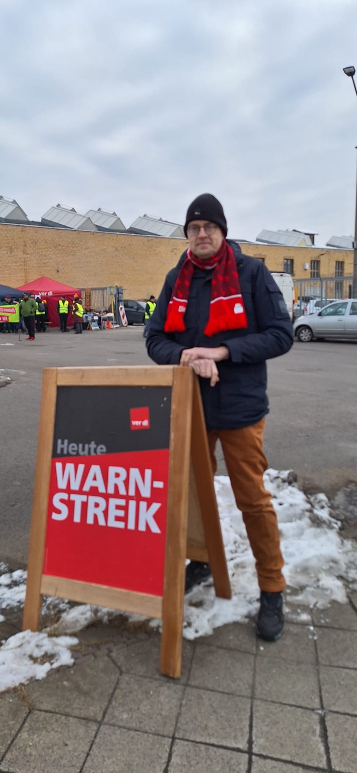 Sören Pellmann steht neben einem Aufsteller: Warnstreik. Im Hintergrund beschäftigte.