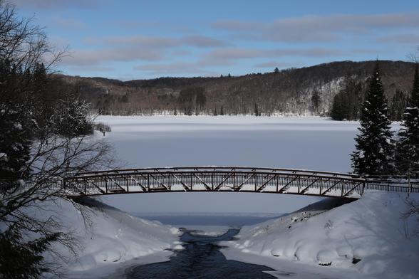 This is a landscape format photo of a winter scene overlooking a frozen lake. The lake empties into a small river in the centre foreground of the photo. The water is not frozen over at this location. An arched  footbridge can be seen extending over the snow covered banks of the river. The lake in the distance is completely frozen and snow covered. A mix of deciduous and coniferous trees can be seen on the opposite shore of the lake over a hilly terrain. A cross country skier can be seen making their way around the lake on the left side of the photo.