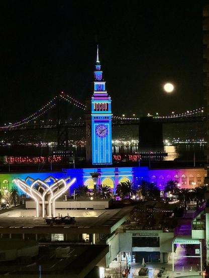 View East from the San Francisco Financial District towards the Ferry Building and Bay Bridge - Ferry building and clock tower are lit up in multiple colors. The moon is full over the bridge and reflecting off the bay.