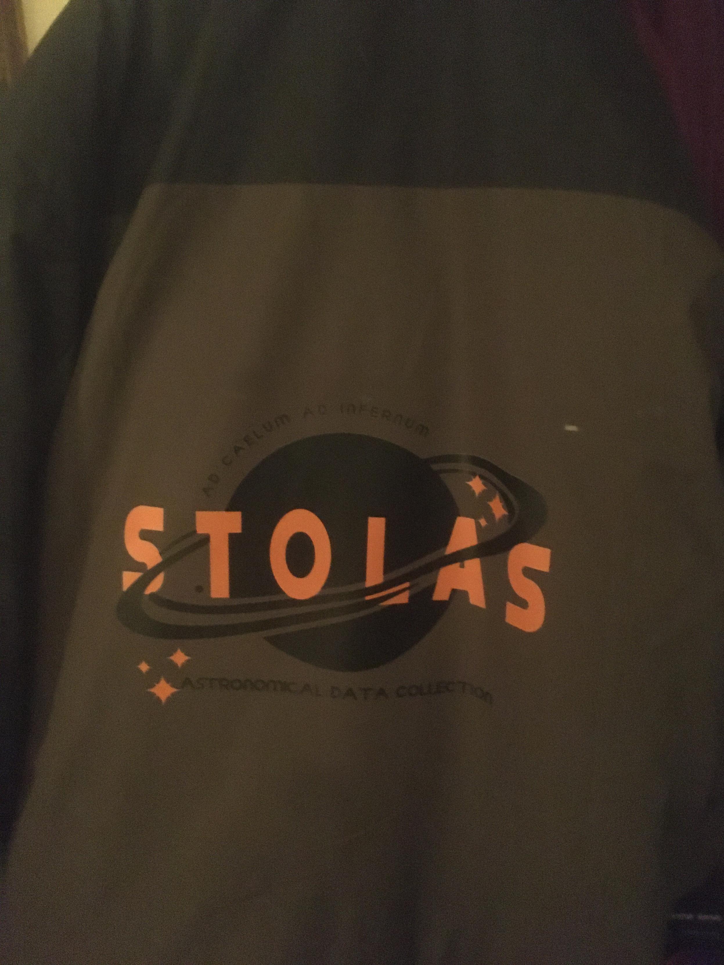 A close-up of a green and black coat, similar to the one Kel wears. A STOLAS logo has been printed on the back of it.