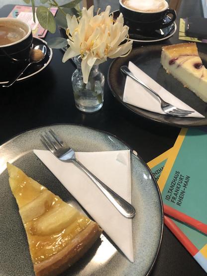 A café table set with two cups of coffee and slices of cake on plates. A small glass vase with a pale yellow flower sits at the center, while colorful ID_Tanzhaus Frankfurt Rhein-Main brochures subtly appear beneath the tableware.